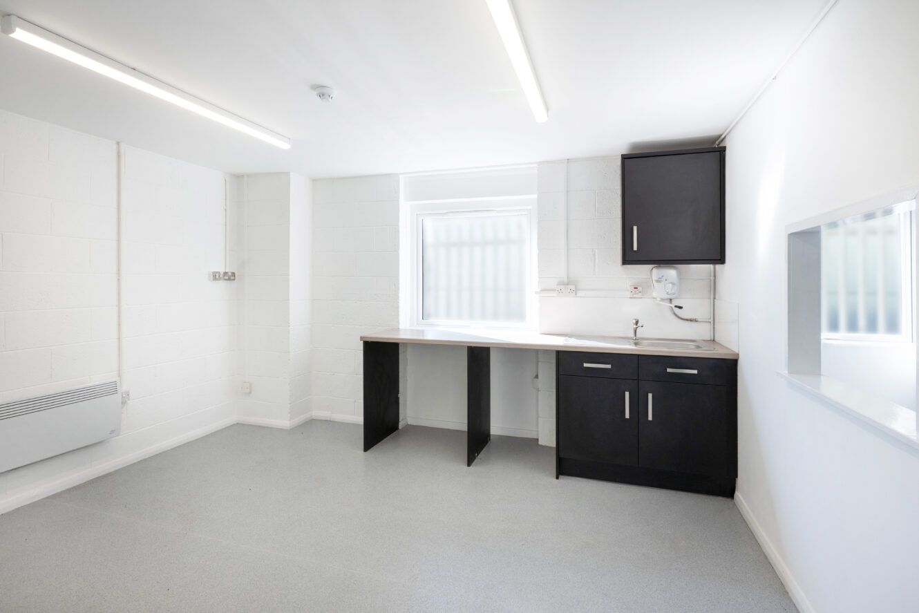 Bright, empty room with white walls, a window, grey flooring, and a kitchenette area with black cabinets and a sink.