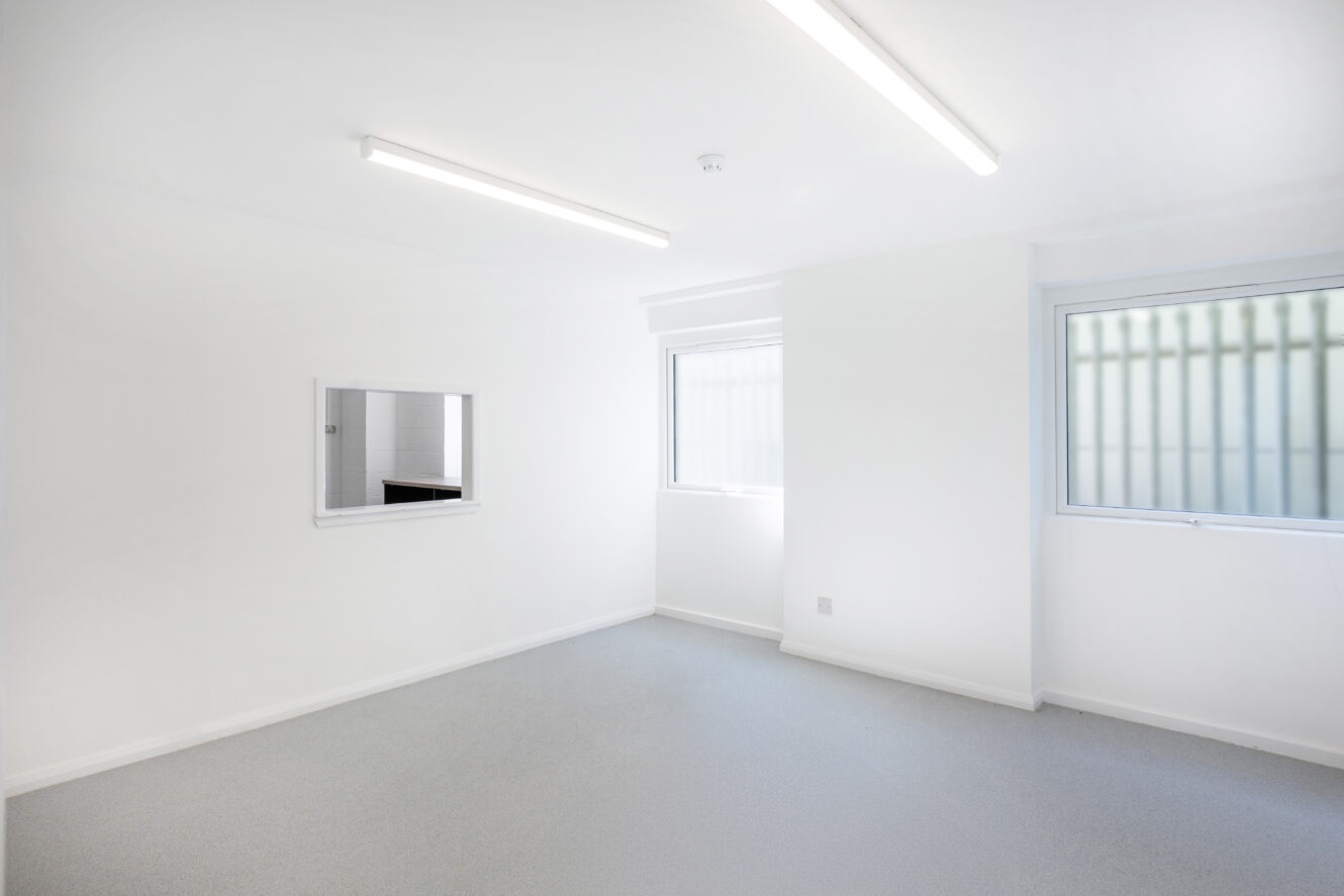 A clean, empty white room with two fluorescent lights, a small window, a larger window, and a wall-mounted pass-through hatch.