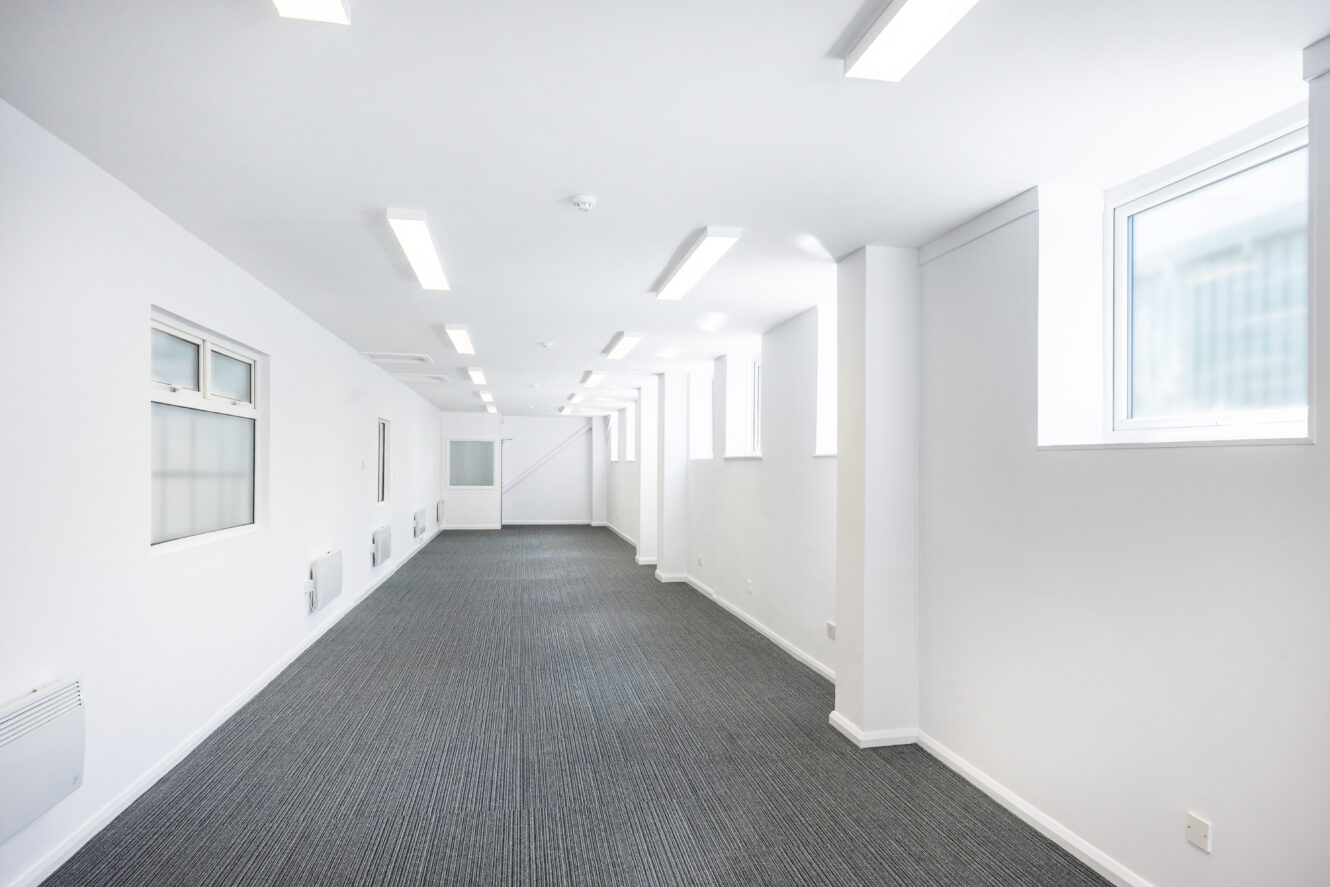 A long, empty office space with white walls, ceiling lights, carpeted floor, and several windows letting in natural light.