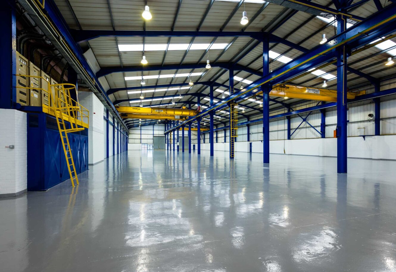 Interior of a large, empty industrial warehouse with blue steel beams, skylights, yellow railings, and polished concrete floor.