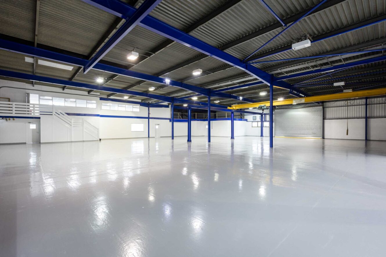 Interior of a large empty warehouse with polished floors, blue support beams, white walls, and overhead lighting. A staircase leads to a mezzanine area on the left.