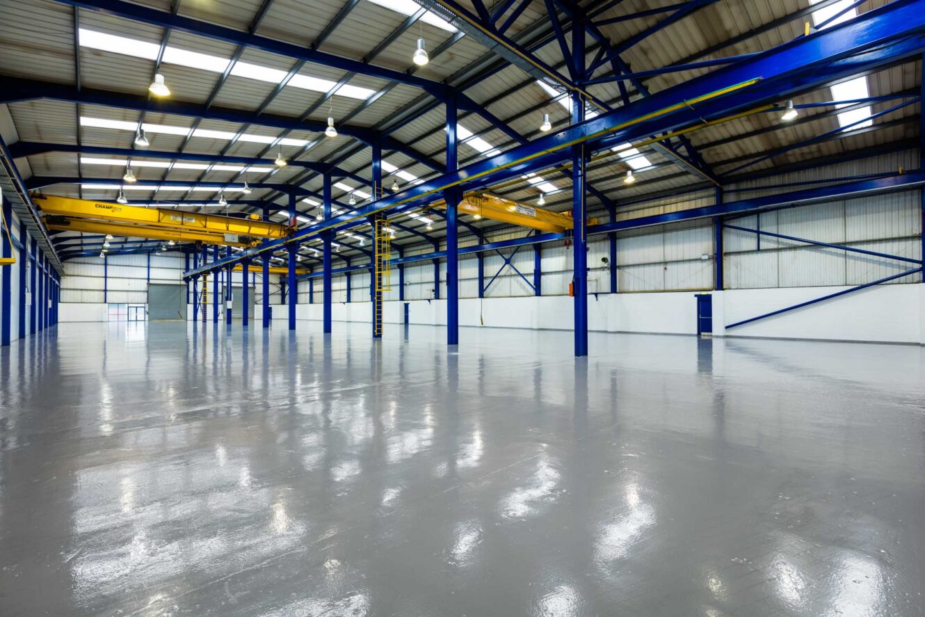Spacious, empty industrial warehouse with polished concrete floors, high ceilings, blue steel beams, and yellow overhead cranes.