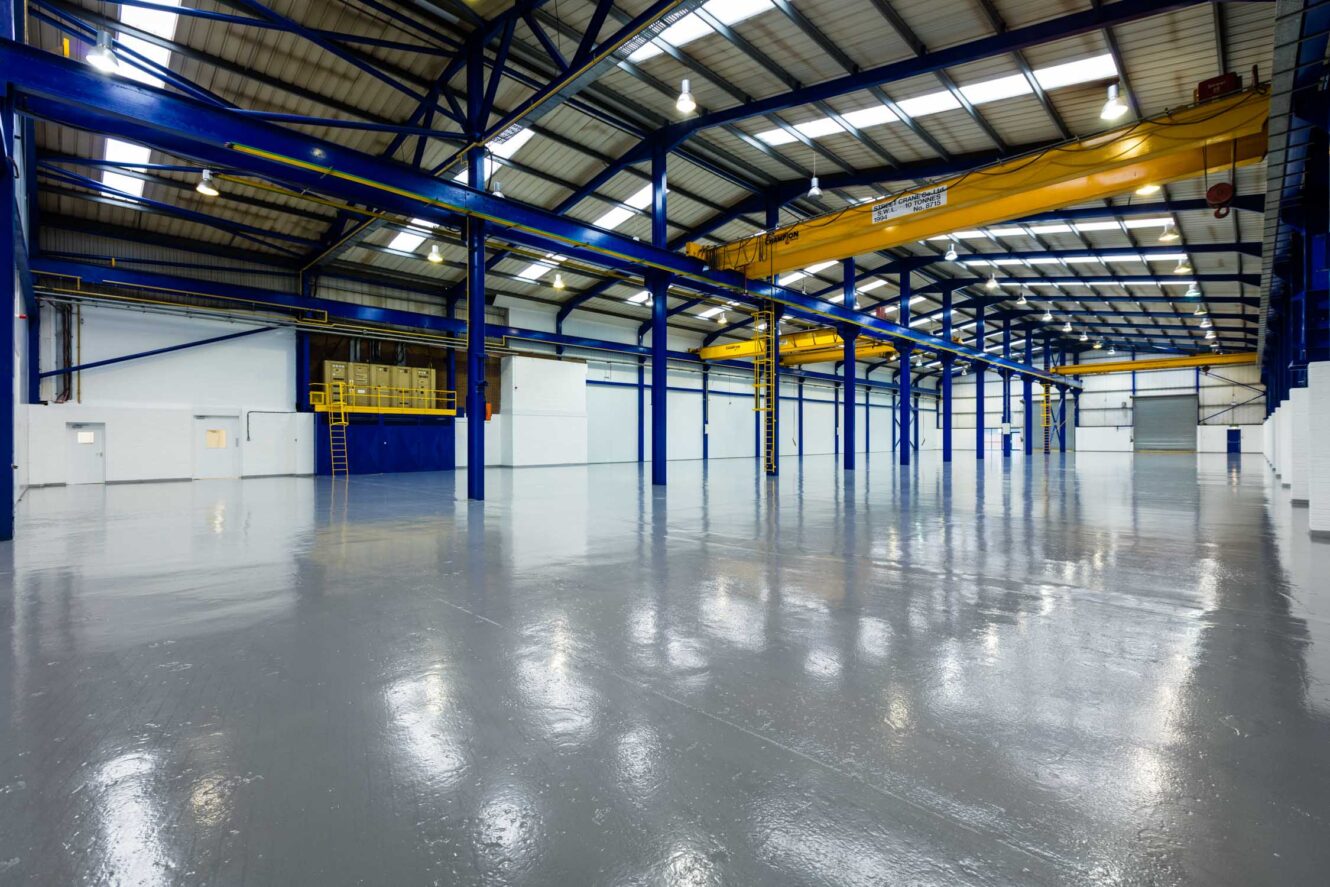 Large, clean, empty industrial warehouse with blue steel beams, yellow cranes, high ceilings, and polished concrete floors. Bright lighting from overhead fixtures.