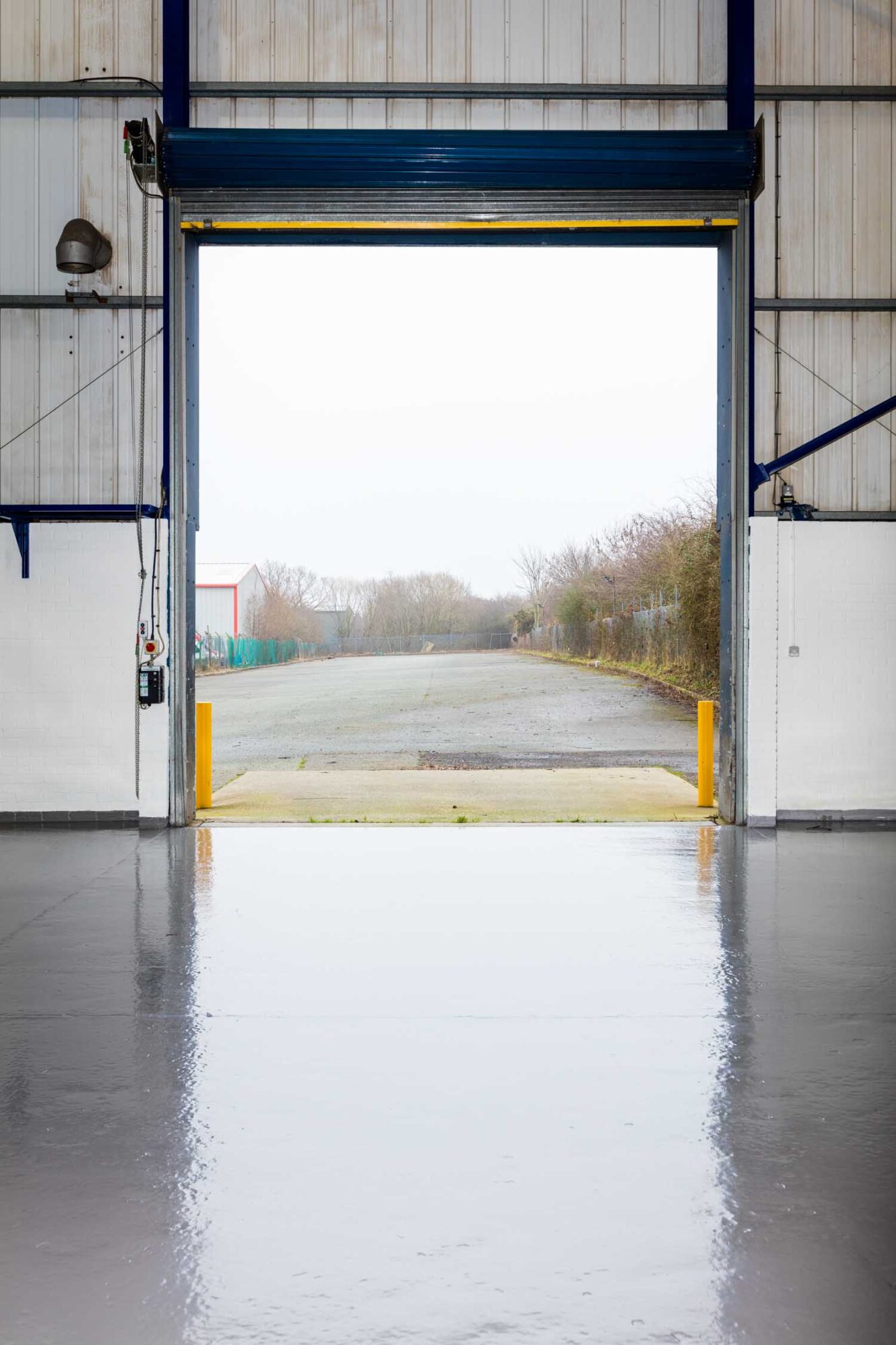 A clean, empty warehouse with an open roller door leading to a paved outdoor area and overcast sky, with trees and fencing visible outside.
