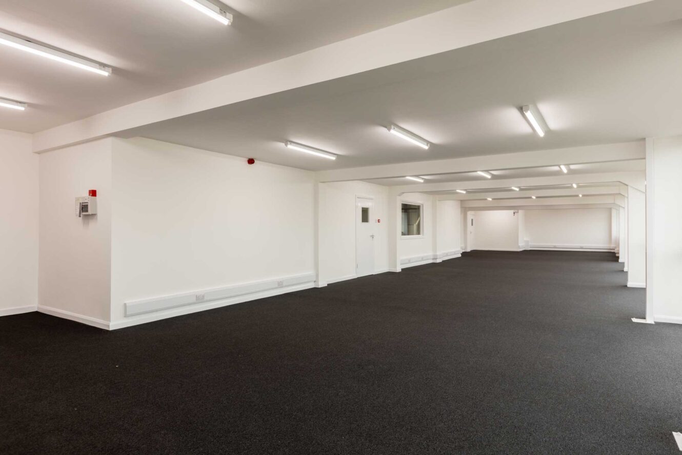 Large, empty office space with white walls, dark carpeted floor, and ceiling-mounted fluorescent lights.