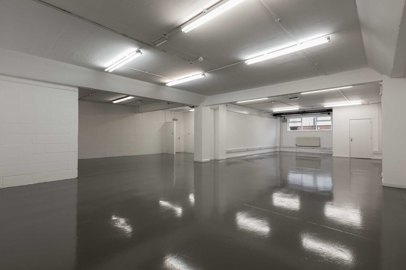 Large, empty room with white walls, bright overhead lighting, and a polished gray floor. Windows and a door are visible at the far end.