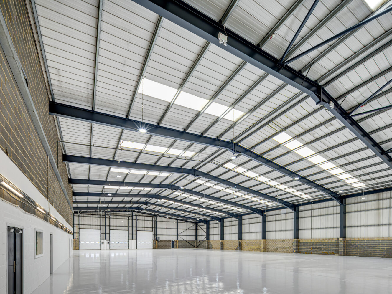 Spacious, empty warehouse interior with high metal ceiling, exposed beams, brick walls, and polished white floor. Large doors line the far wall.