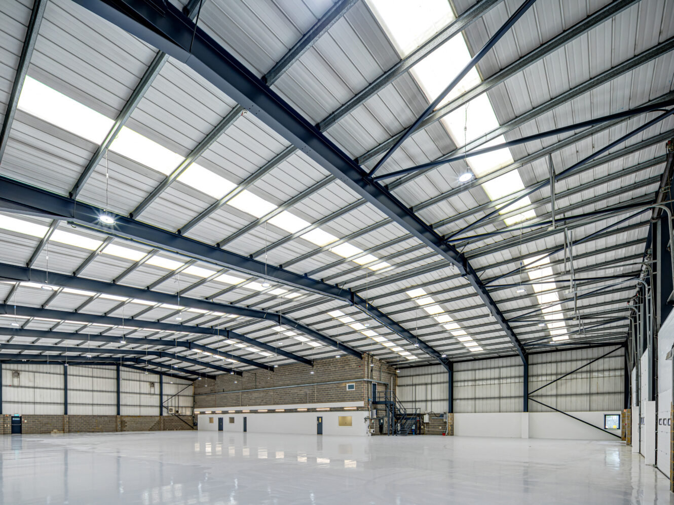 Large empty warehouse with high metal ceiling, white floors, and exposed beams; brick and paneled walls, and a staircase leading to a mezzanine level.