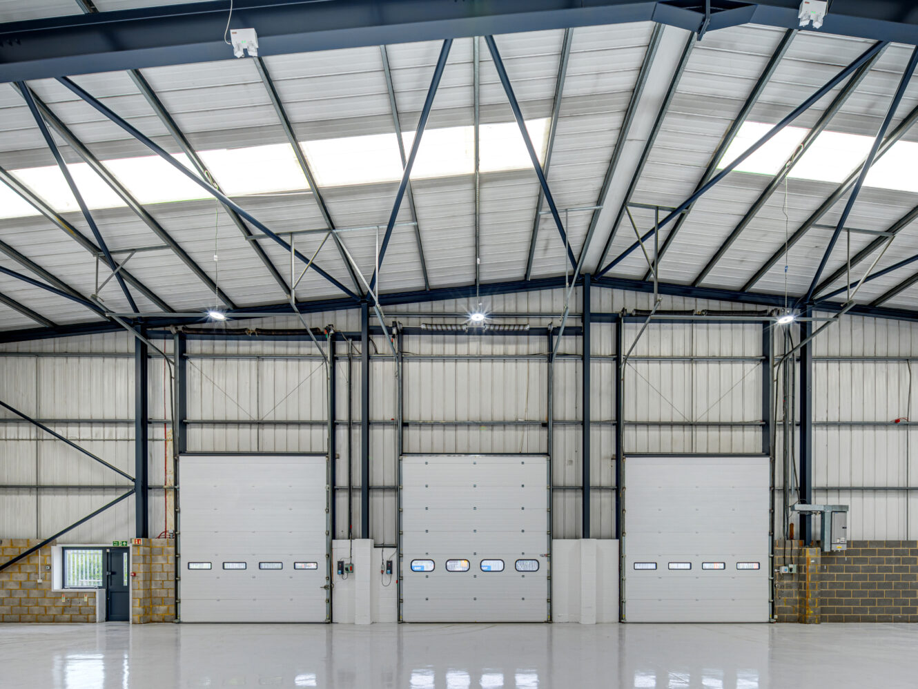 Interior of an empty industrial warehouse with high ceilings, metal beams, and three large white loading bay doors.