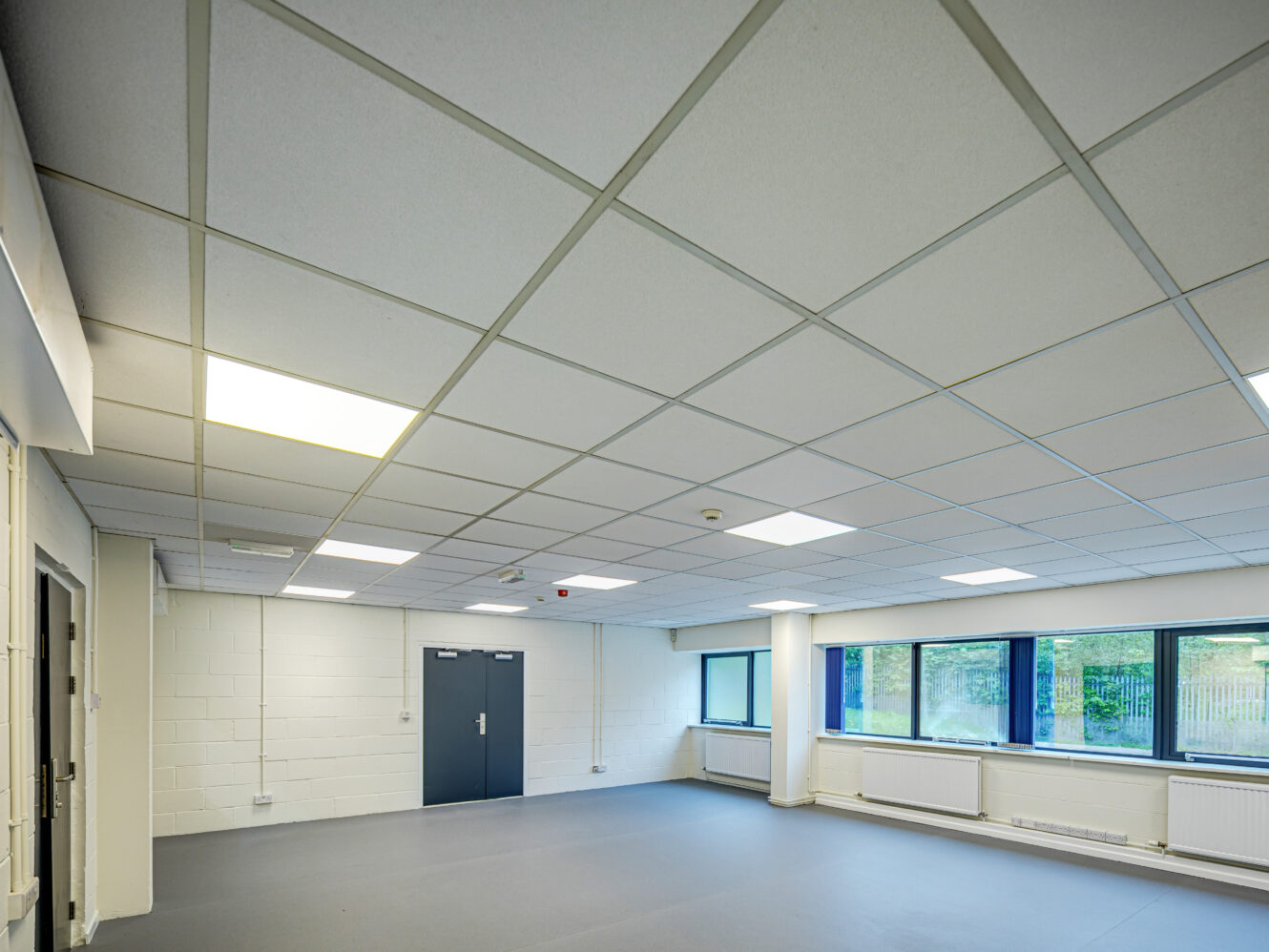 Empty room with a suspended ceiling, fluorescent lighting, white brick walls, large windows, gray double doors, and a bare gray floor.