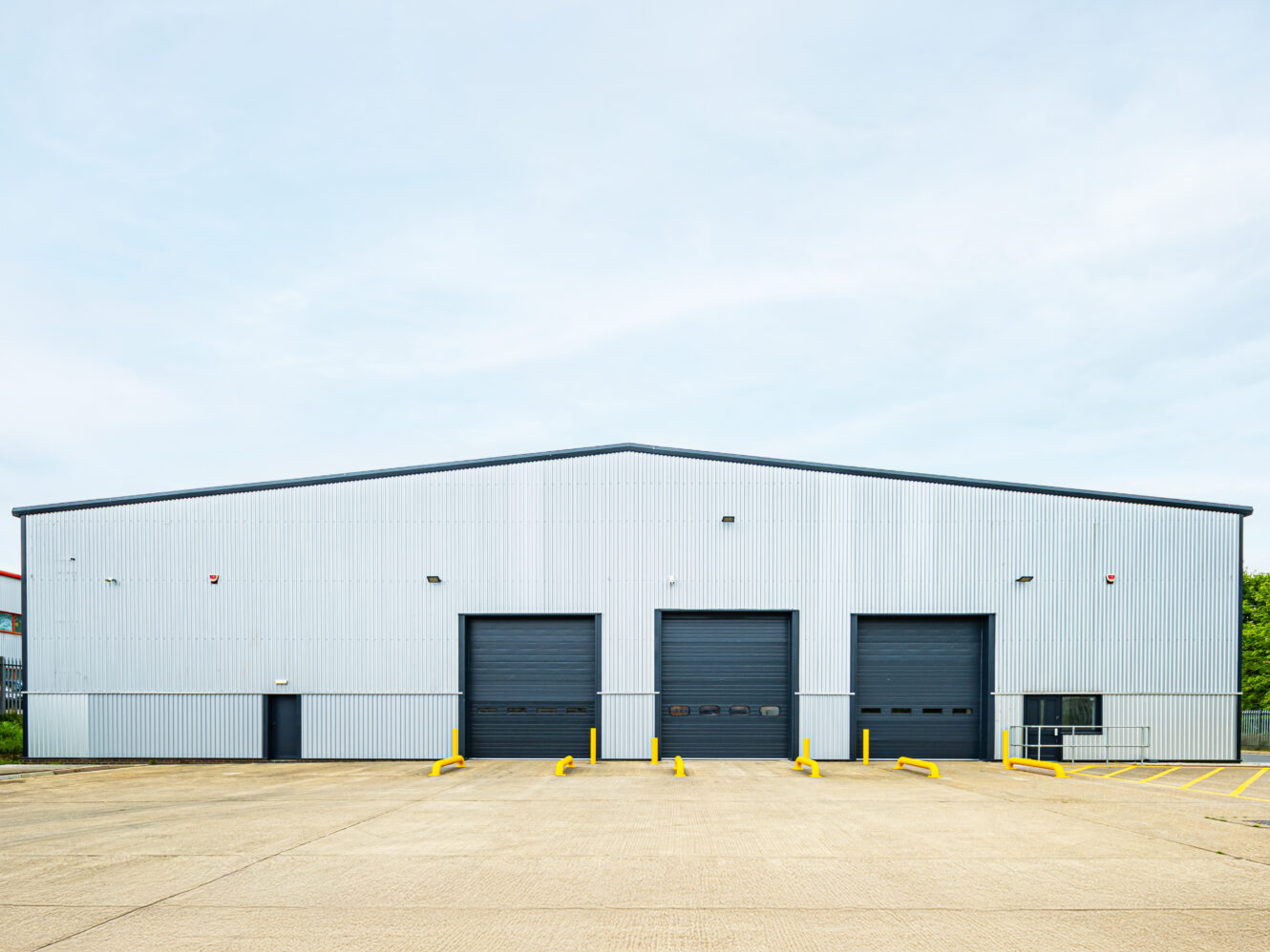 A large industrial warehouse with three black loading bay doors, a gray metal exterior, yellow safety barriers, and an empty concrete parking lot.