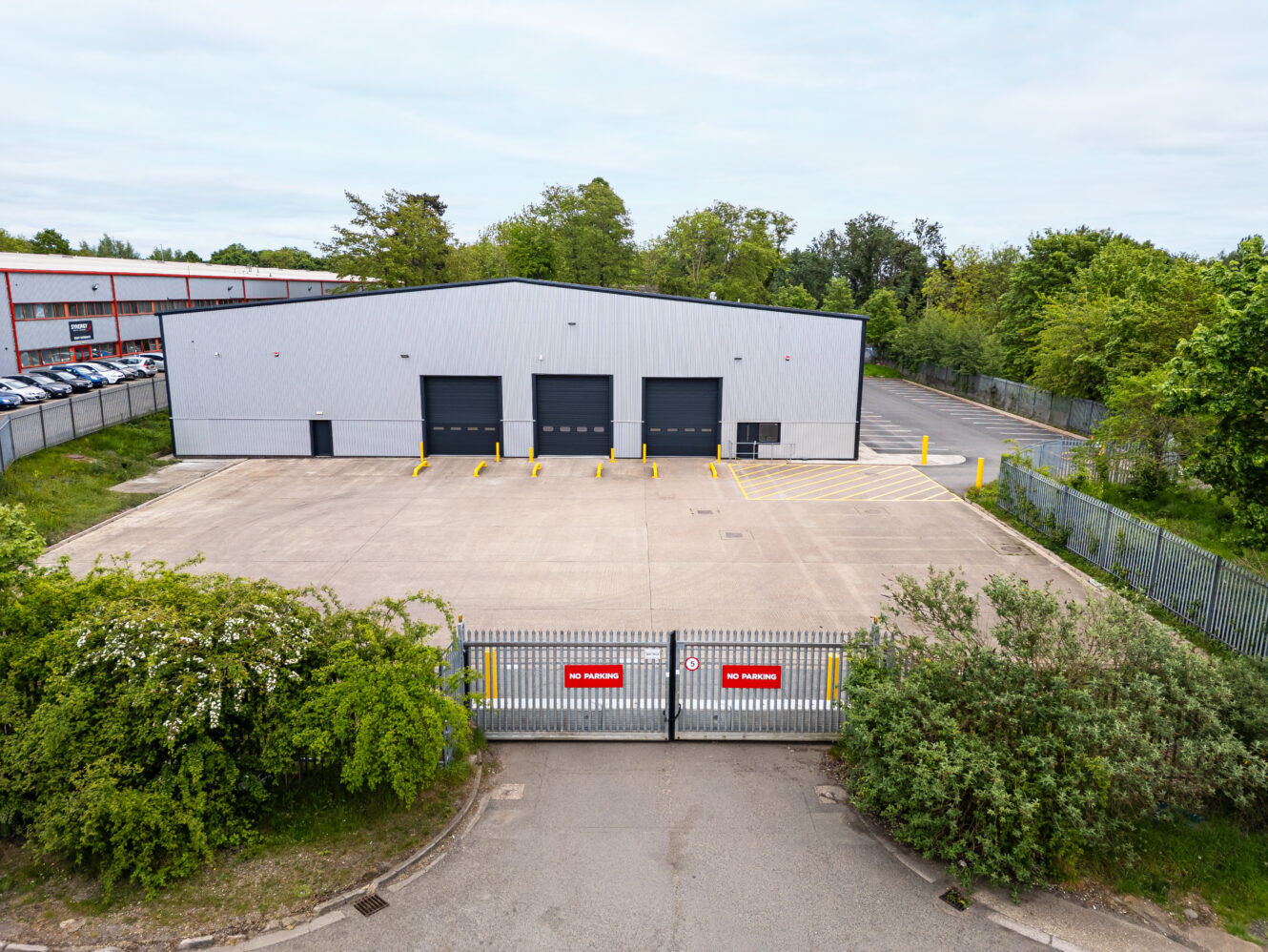 A large, empty industrial warehouse with three loading bays and a fenced, gated entrance displaying No Parking signs. Surrounding area features trees and parked cars in the background.