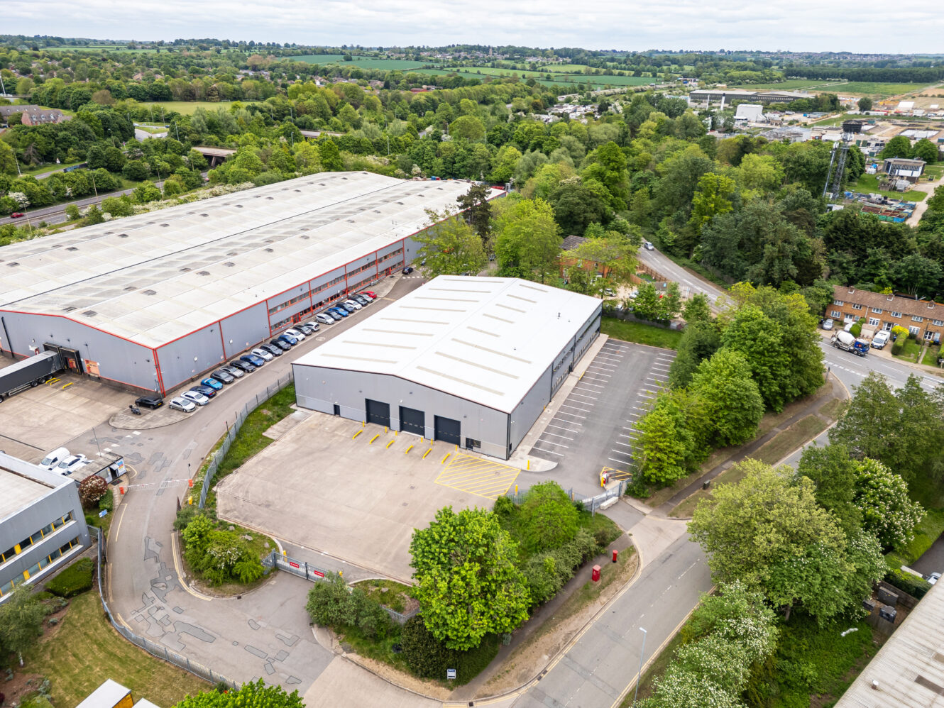 Aerial view of a large industrial warehouse with a parking lot, surrounded by trees and roads, and adjacent to another larger warehouse.