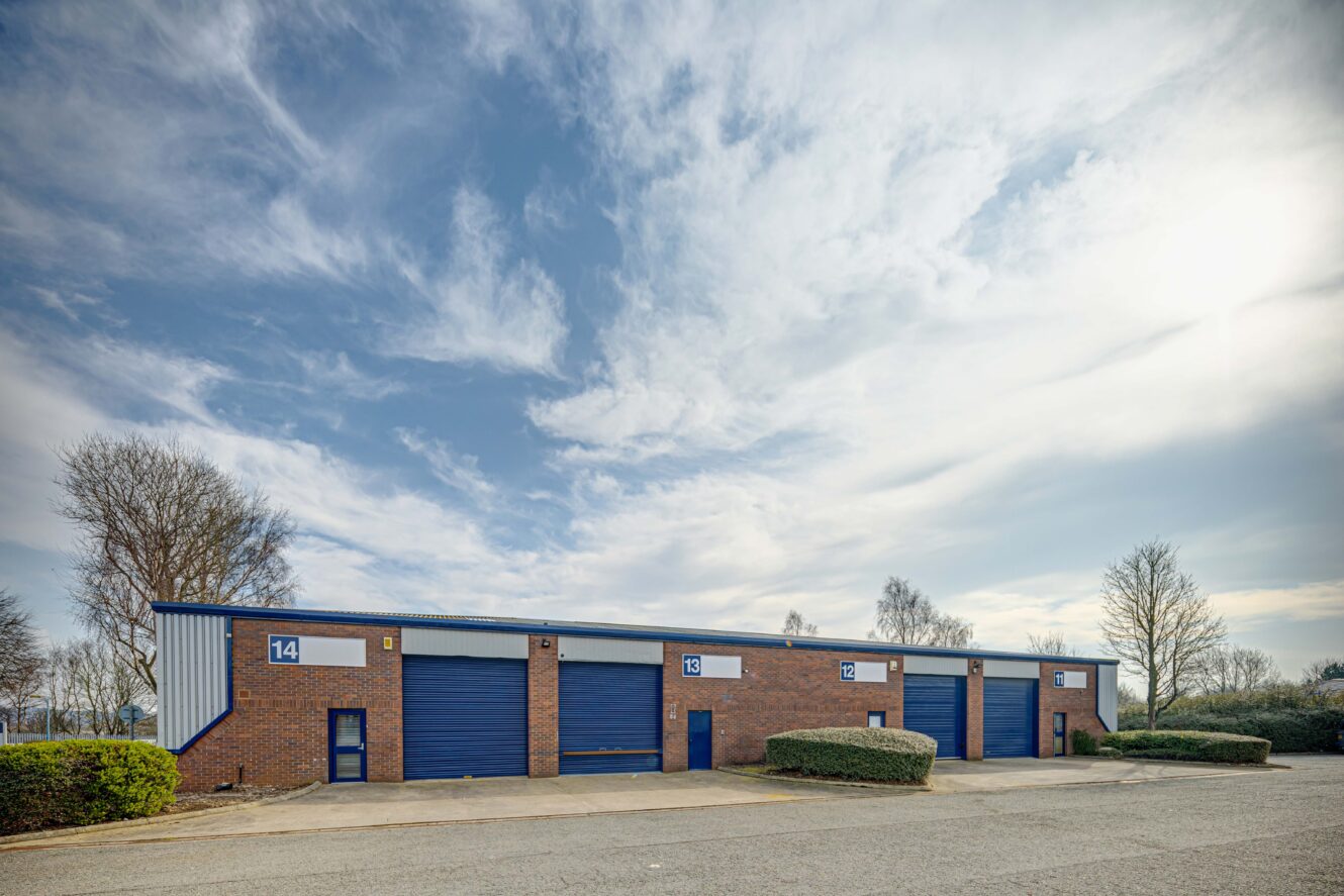 A row of brick industrial warehouse units with blue doors and numbered signs, surrounded by bushes and trees under a partly cloudy sky.