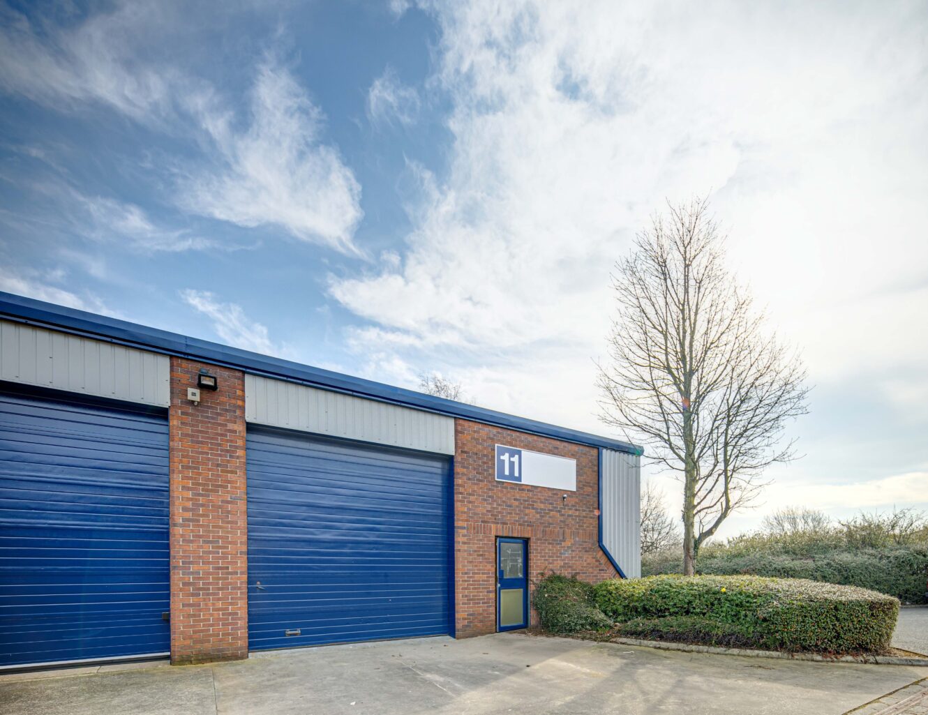 A warehouse unit with blue roller doors and a small entry door marked with the number 11, next to a leafless tree and shrubbery under a partly cloudy sky.