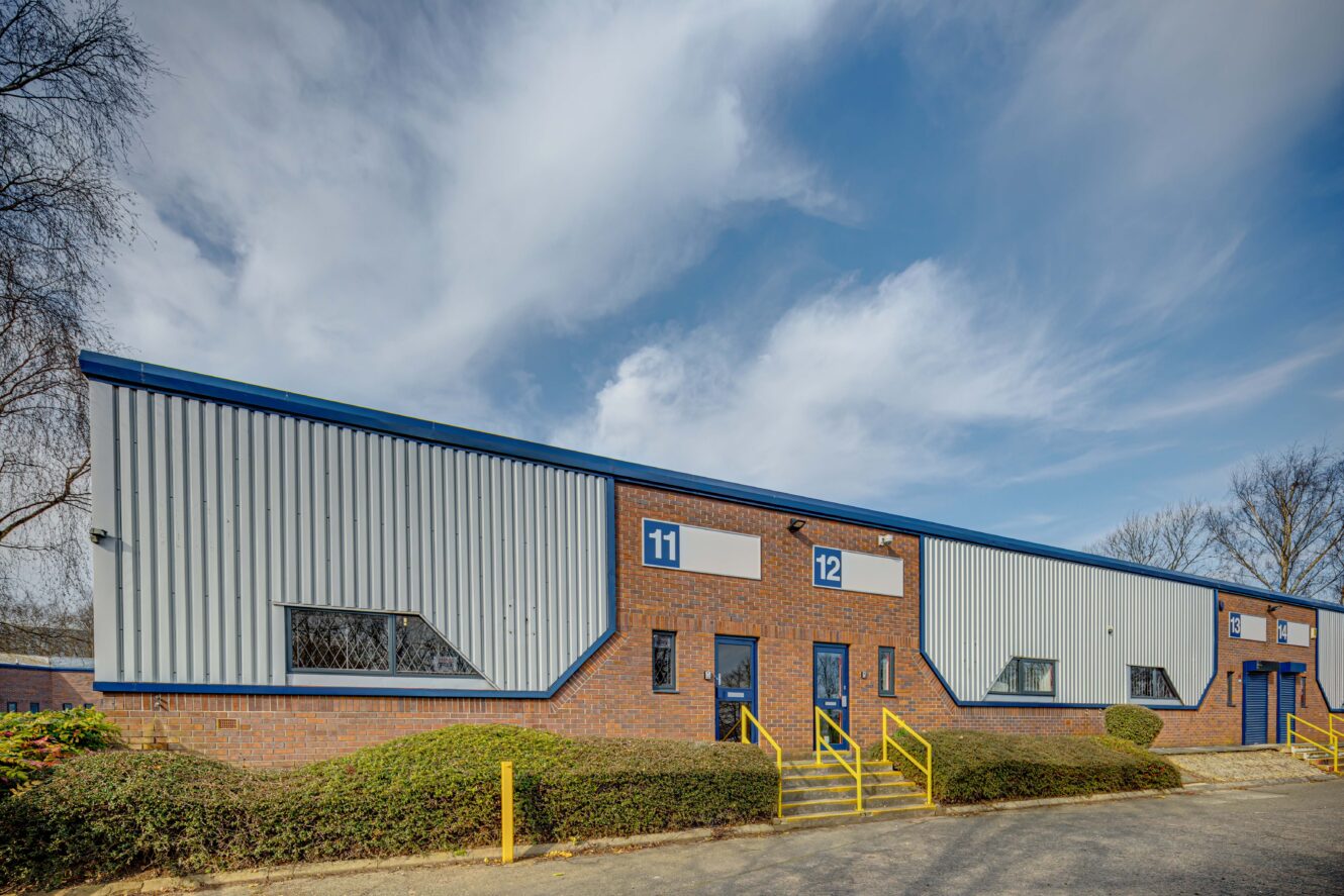 A single-story industrial building with blue and white trim, labeled units 11 and 12, yellow railings, brick facade, and a cloudy sky overhead.