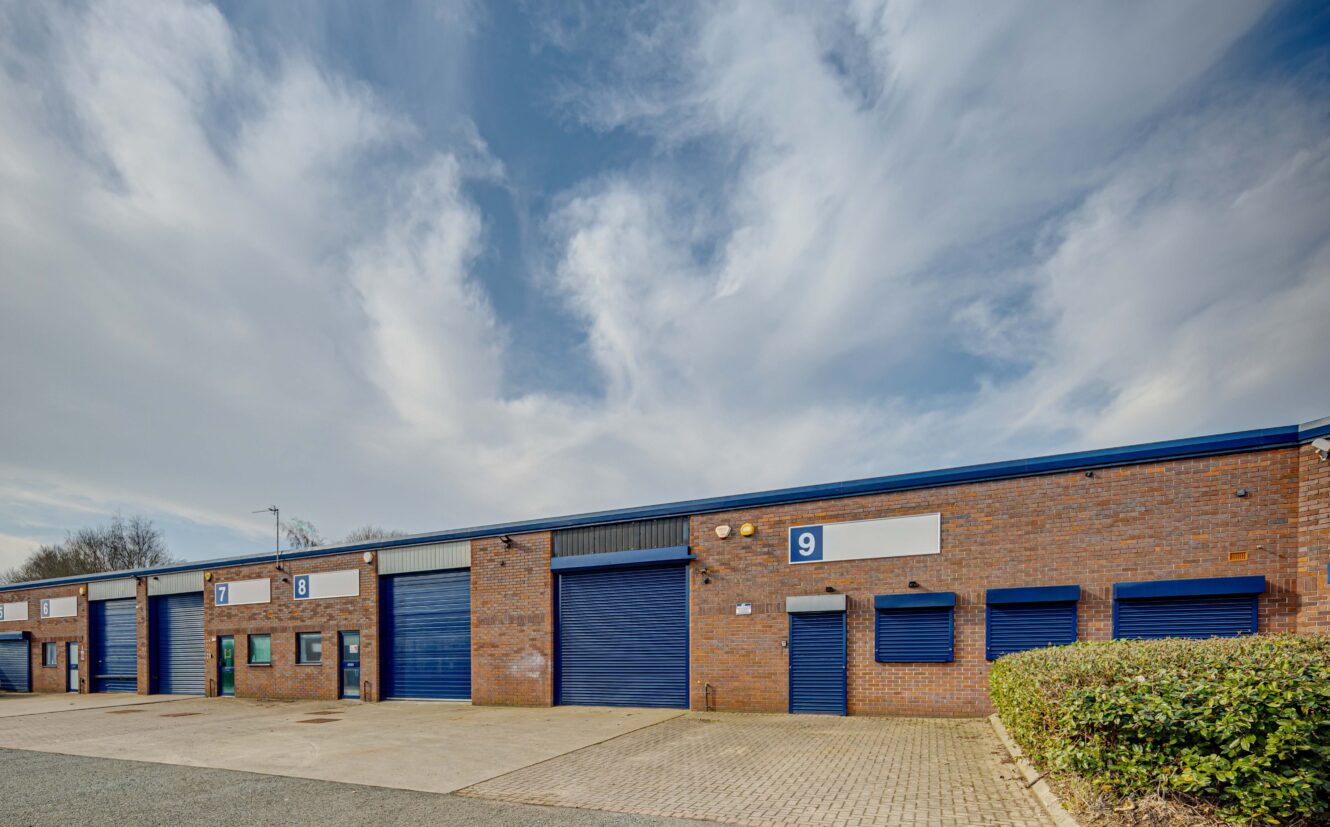 A row of industrial units with blue roller doors and brick walls, labeled with numbers including 9, under a partly cloudy sky.