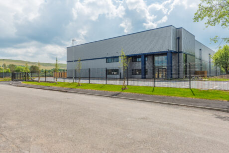 A modern, grey industrial warehouse with large windows, surrounded by a black metal fence and young trees, sits next to an empty road under a partly cloudy sky.