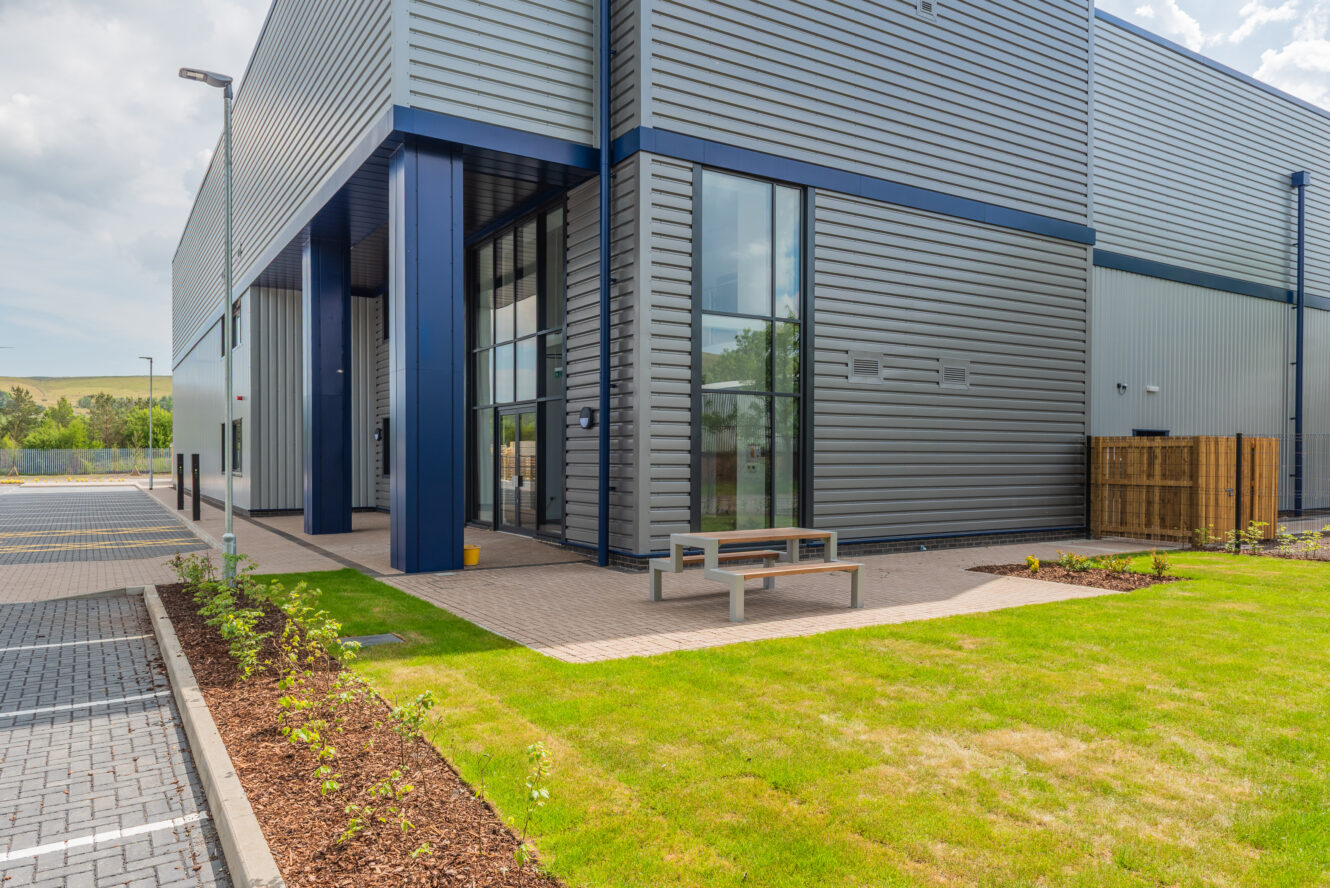 Modern industrial building with large windows, metal siding, and a landscaped area featuring grass, shrubs, and a bench near a paved walkway and parking spaces.