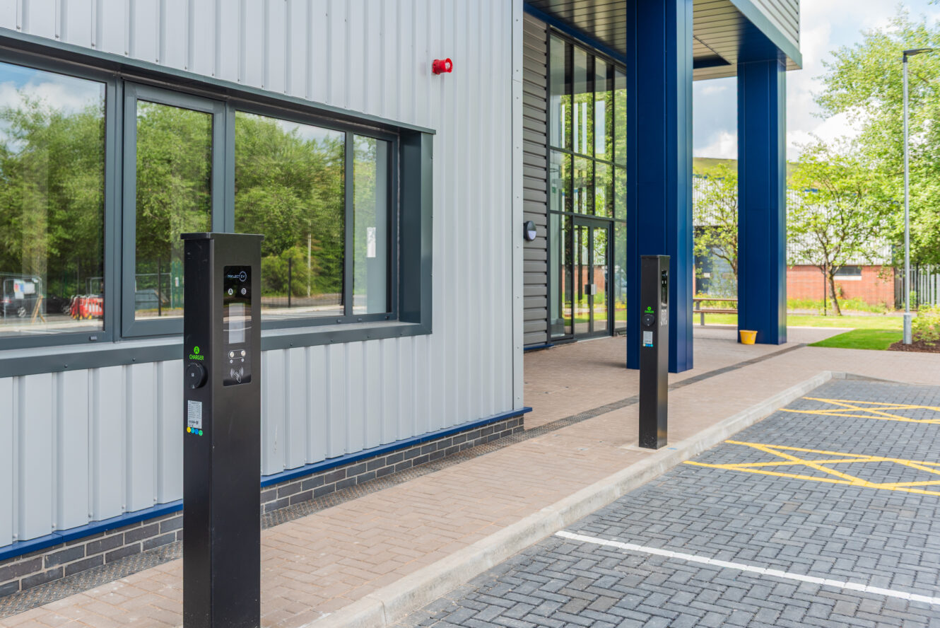 Two electric vehicle charging stations are installed in front of a modern commercial building with large windows and blue columns.