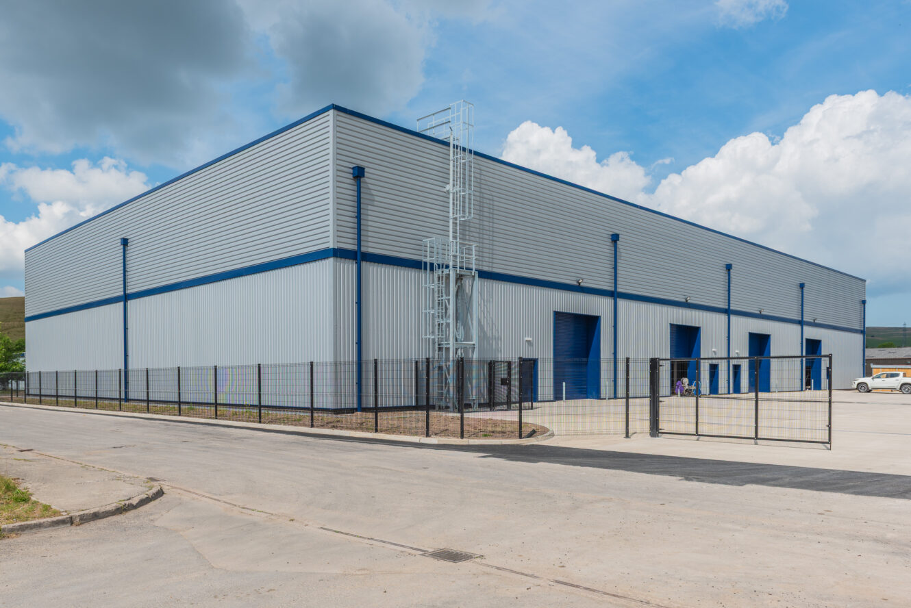 A large, modern industrial warehouse with metal siding, blue accents, several loading doors, and a fenced perimeter under a partly cloudy sky.