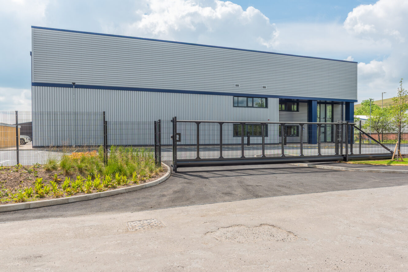 A modern industrial building with metal cladding, large windows, and a closed security gate in front, surrounded by fencing and landscaping.