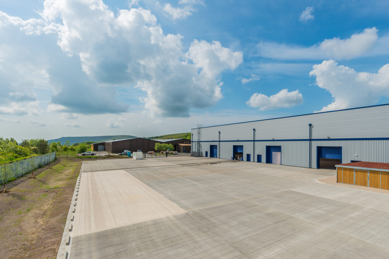 Large industrial warehouse with multiple blue loading bays and a spacious concrete lot under a partly cloudy sky. Trees and a smaller building are visible in the background.