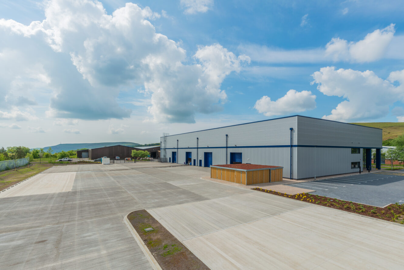 A large industrial warehouse with blue doors and a spacious concrete parking area under a partly cloudy sky.