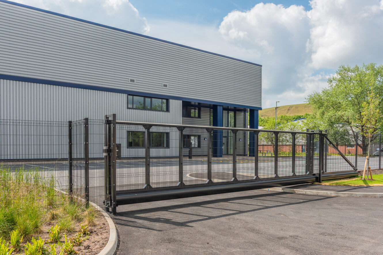 A sliding security gate is closed in front of a modern industrial building with metal siding and large windows.