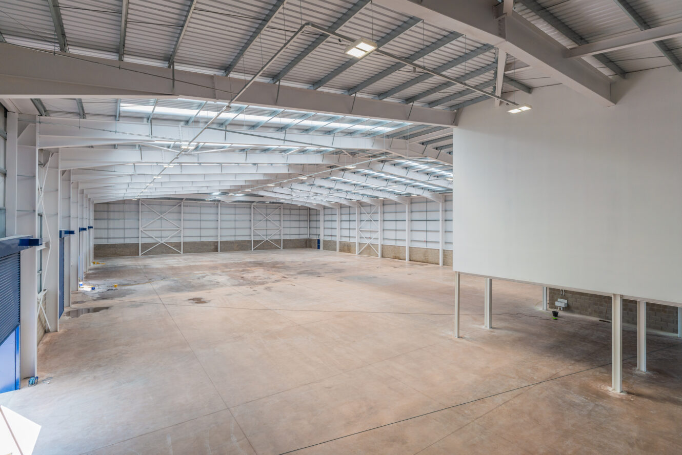Large, empty industrial warehouse with high ceilings, exposed beams, concrete floor, and ample overhead lighting.
