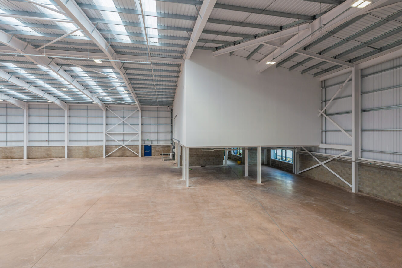 Empty industrial warehouse interior with high ceilings, large skylights, concrete floor, and a white mezzanine office space on metal stilts.