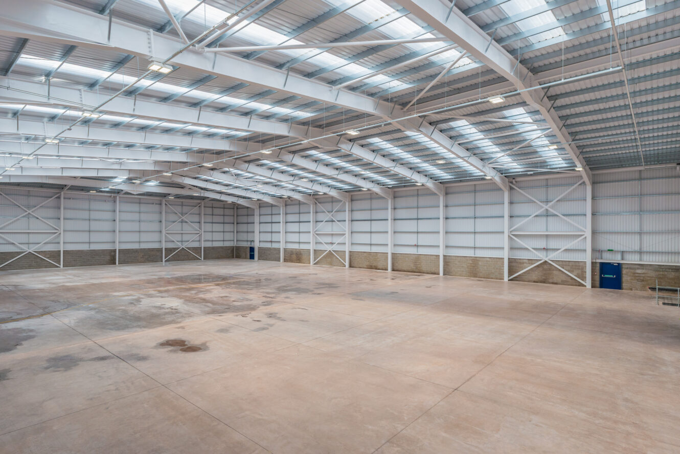 Spacious empty warehouse interior with high ceilings, metal beams, concrete floor, and skylights allowing natural light inside.