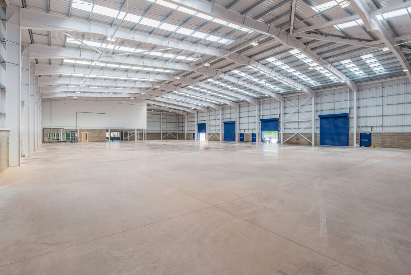 Large empty warehouse with high ceilings, concrete floor, metal beams, skylights, and several blue roll-up doors along one wall.