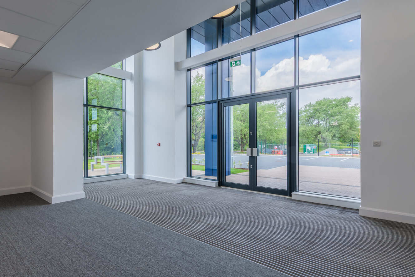 A modern, empty lobby with large floor-to-ceiling windows, gray carpet, and double glass doors leading outside to a paved area and greenery.