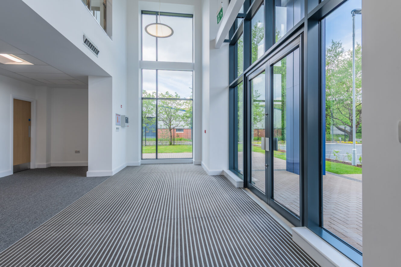 Modern building interior with large windows, glass doors, and striped carpet leading to an outdoor area with trees and brick paving.