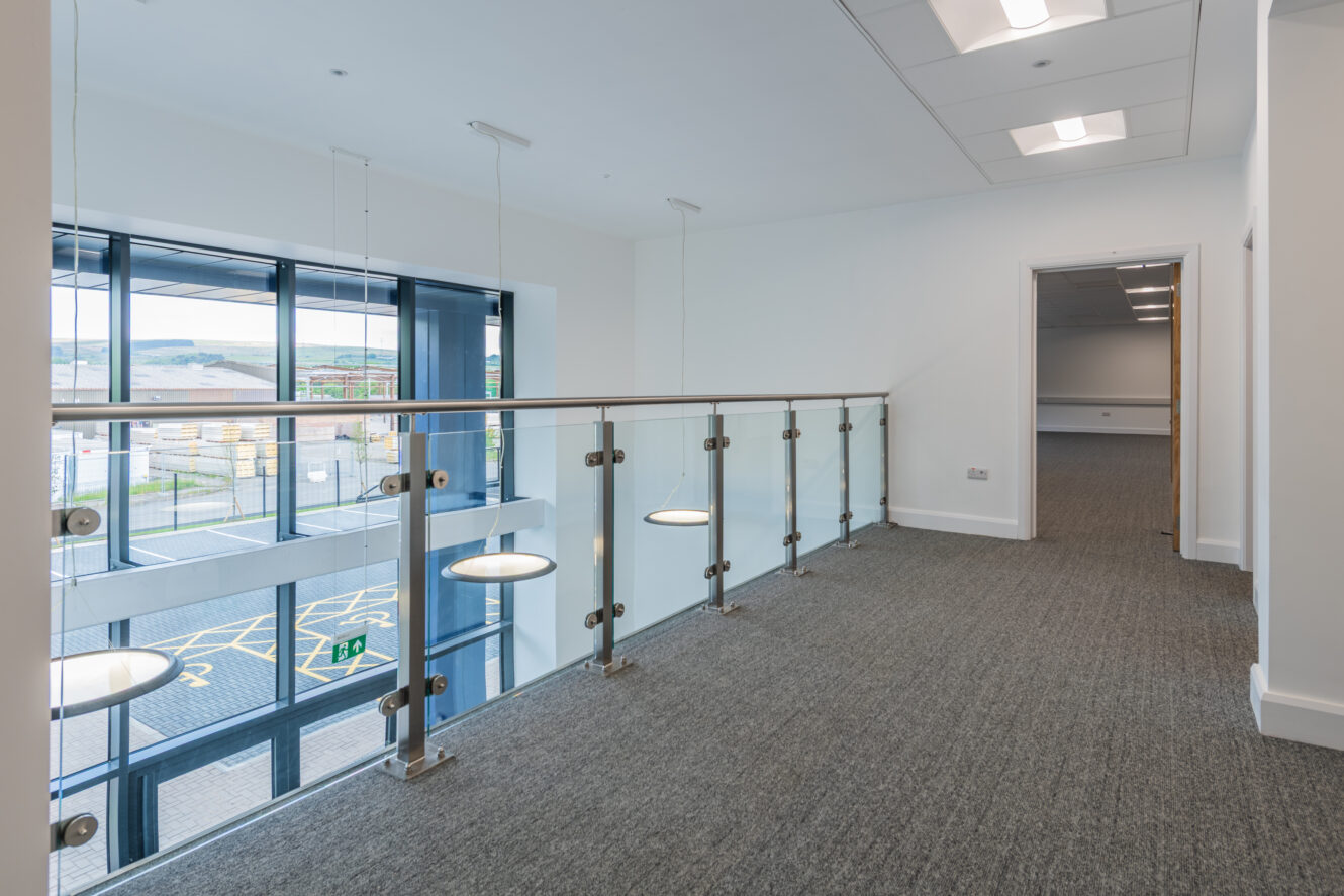 Modern office interior with glass balcony railing overlooking parking lot, grey carpet flooring, white walls, and LED ceiling lights; open doorway leads to another empty room.