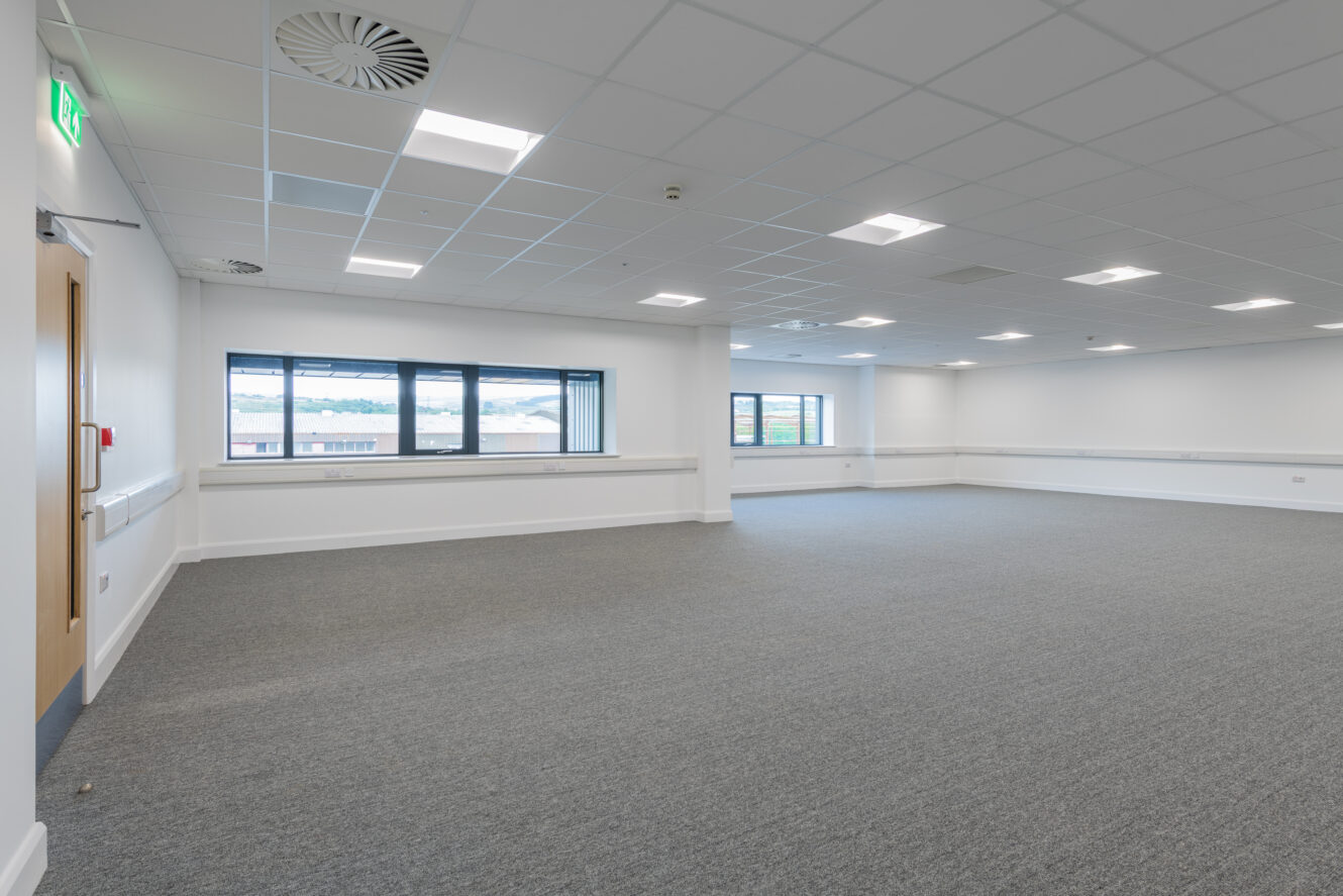 Spacious, empty room with gray carpet, white walls, large windows, ceiling lights, and a closed wooden door.