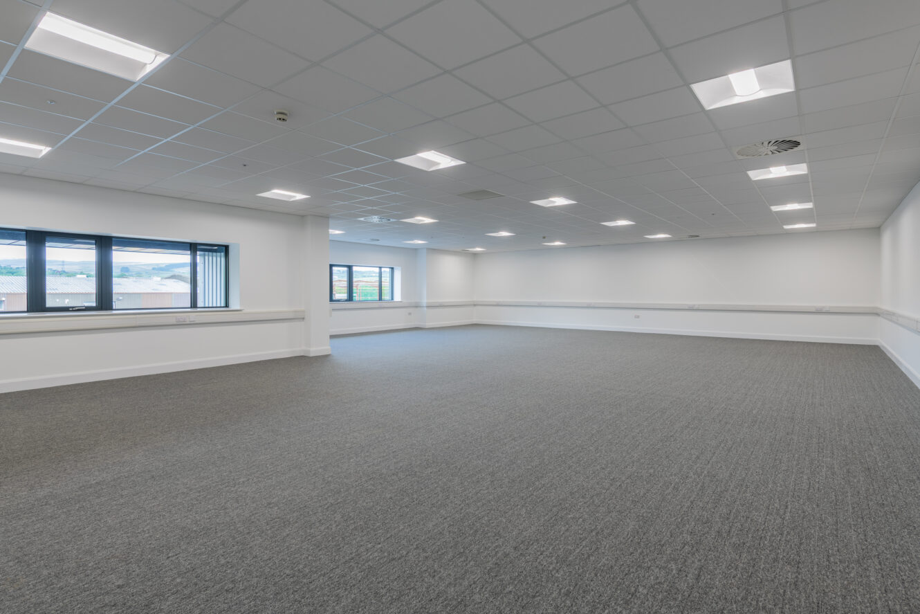 Empty, modern office room with gray carpet, white walls, drop ceiling, fluorescent lighting, and large windows along one wall.