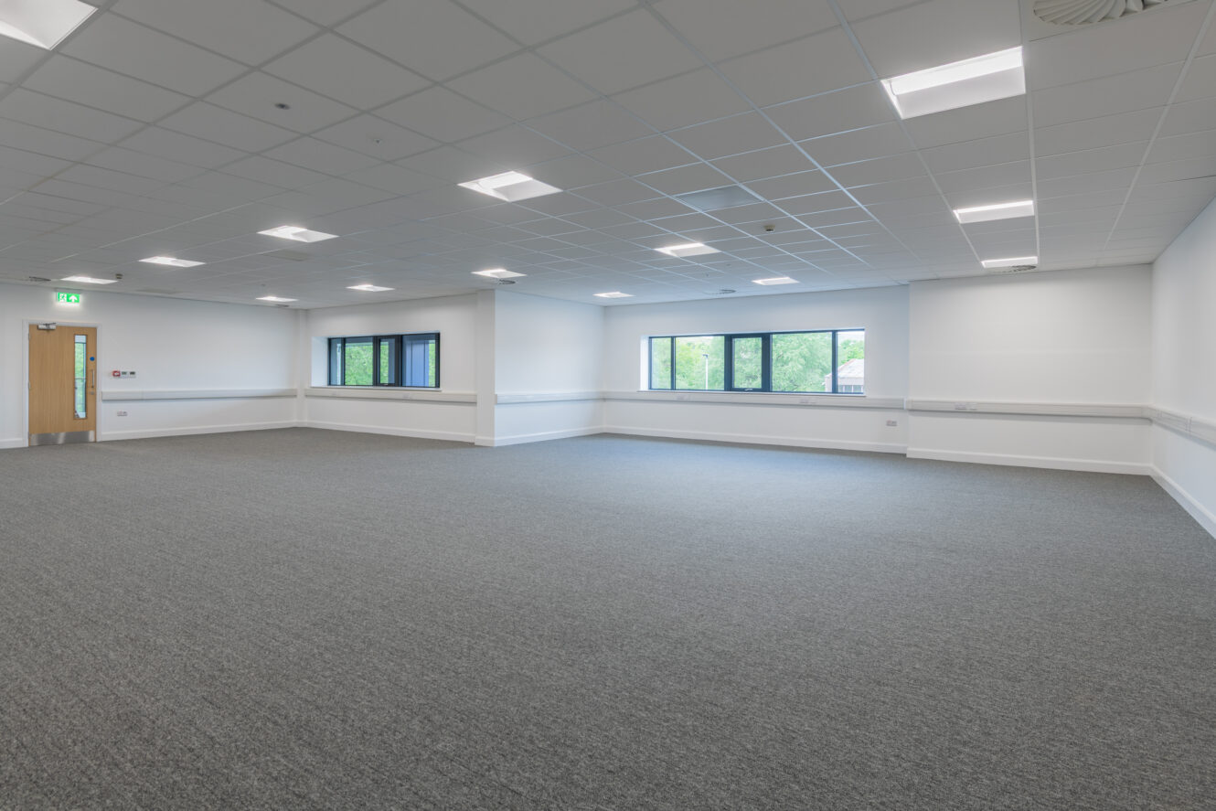 A large, empty room with carpeted floor, white walls, ceiling lights, multiple windows, and a closed wooden door.