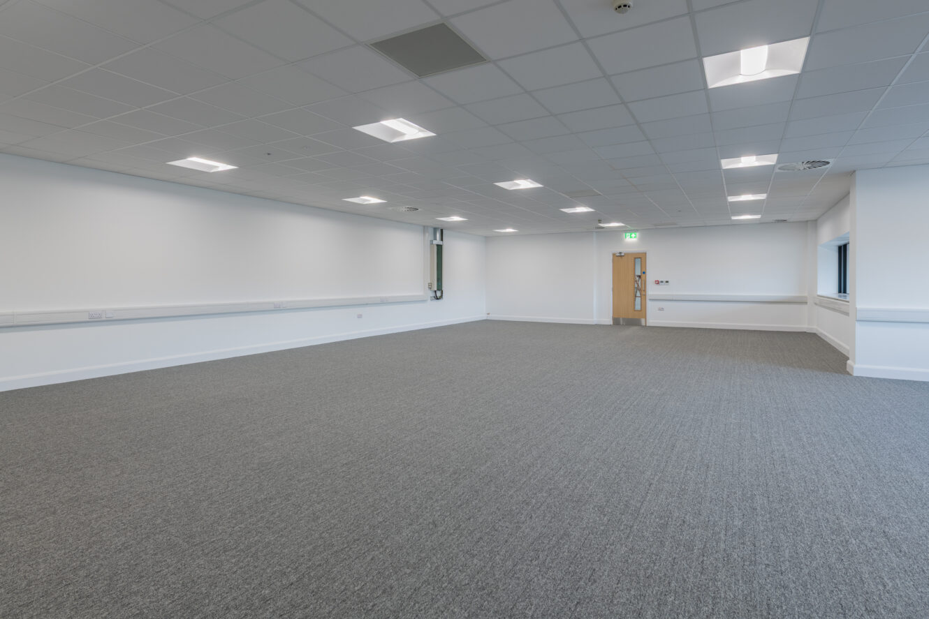 Large empty office space with grey carpet, white walls, a suspended ceiling with fluorescent lighting, and a closed wooden door at the far end.