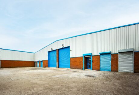 A warehouse building with white walls, blue doors, and loading bays numbered 5 and 6, set in an empty paved lot under a clear sky.