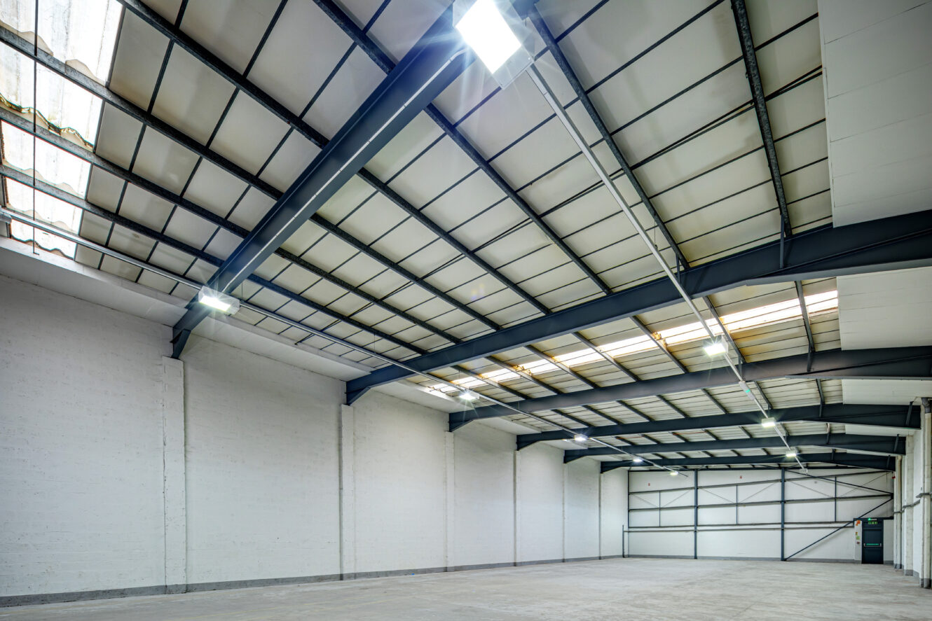 Interior of an empty industrial warehouse with high ceilings, exposed steel beams, white walls, and large overhead lights.