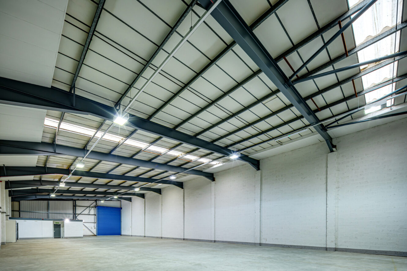 Spacious empty warehouse with high ceiling, exposed steel beams, white brick walls, concrete floor, and blue roller shutter door. Bright lighting fixtures are mounted on the ceiling.