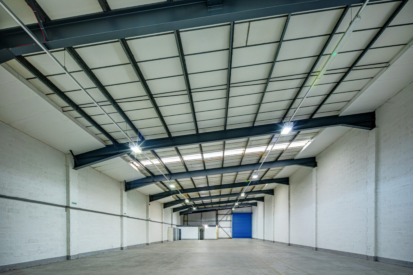 Interior view of a large, empty industrial warehouse with white brick walls, a concrete floor, and a high ceiling featuring skylights and exposed steel beams.