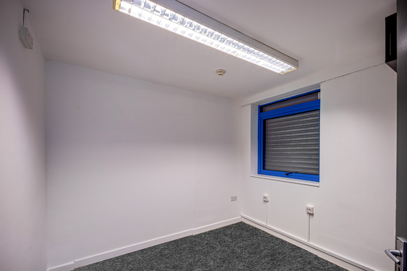 A small, empty room with white walls, a gray carpet, a single blue-framed window with a metal shutter, and a fluorescent ceiling light.