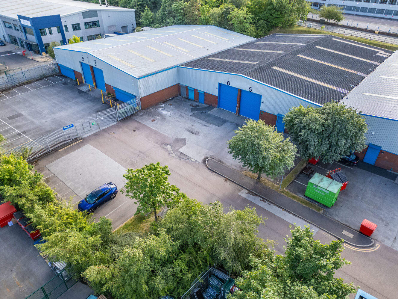 Aerial view of an industrial warehouse complex with blue roller doors, parked cars, trees, and a fenced perimeter on a clear day.