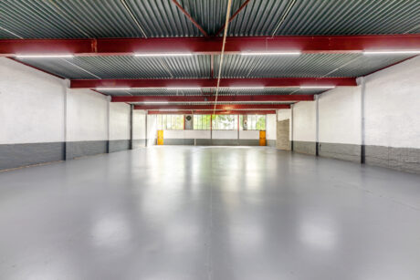 Empty industrial warehouse space with a grey concrete floor, white and grey walls, exposed beams, and large windows along the back wall letting in natural light.