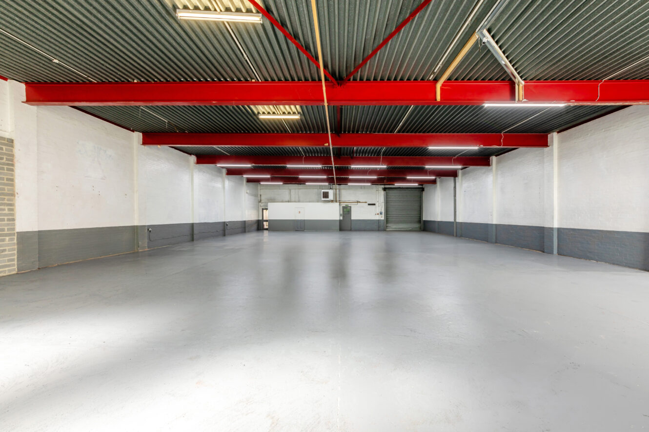 Spacious empty warehouse with gray flooring, white and gray walls, red ceiling beams, and fluorescent lighting. A garage door and entry doors are visible at the far end.