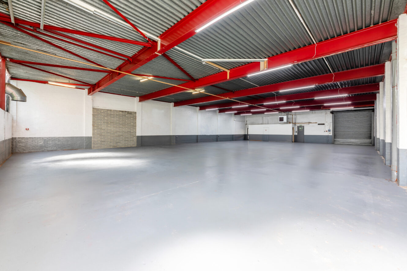 Large empty warehouse interior with gray floor, white and gray walls, red ceiling beams, fluorescent lighting, and two large metal roller doors.