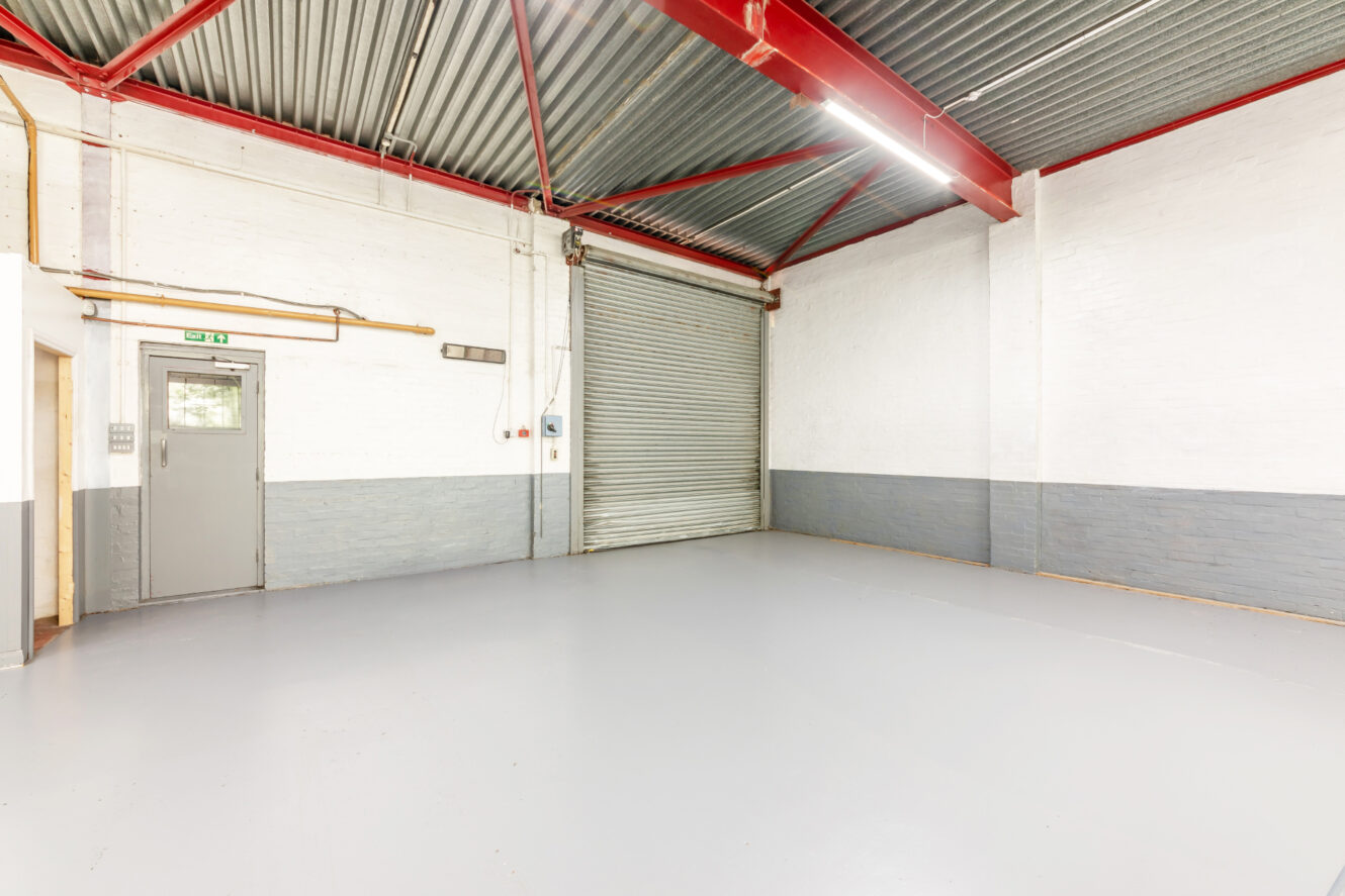 Empty industrial warehouse unit with gray concrete floor, white and gray painted brick walls, a metal roller shutter door, and exposed red steel beams.